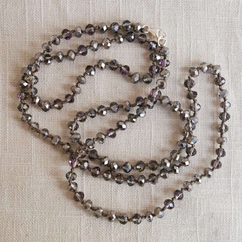 A single-strand, hand-knotted necklace made of faceted aurora borealis crystals is displayed draped on a linen. 