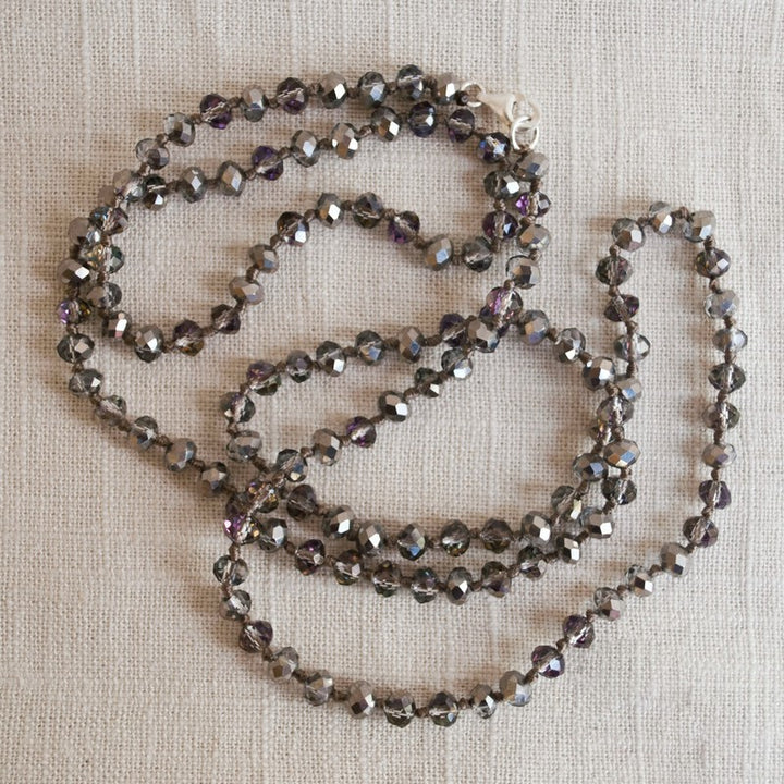A single-strand, hand-knotted necklace made of faceted aurora borealis crystals is displayed draped on a linen. 
