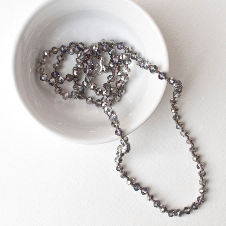 A single-strand, hand-knotted necklace made of faceted aurora borealis crystals is displayed draped on a small white bowl.