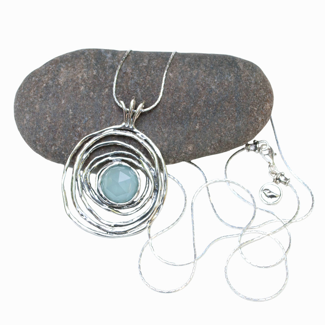 A sterling silver pendant necklace with a central chalcedony stone, displayed on a pebble surface. The necklace is 30 inches long and has a lobster closure.