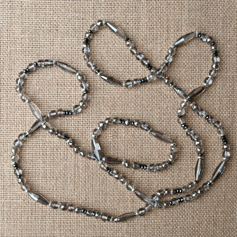 A long necklace with various-shaped crystals punctuated with hematite beads is shown on a linen background.