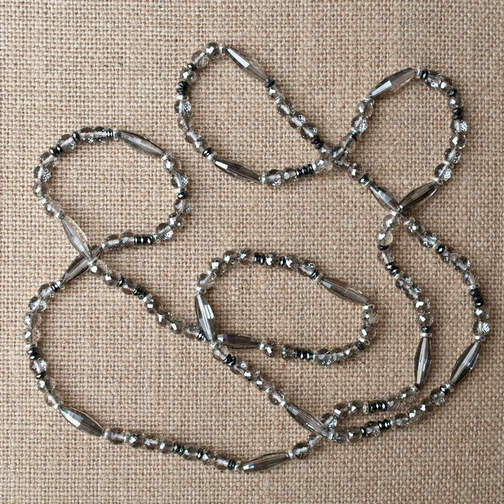 A long necklace with various-shaped crystals punctuated with hematite beads is shown on a linen background.