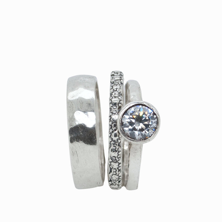 A set of three silver rings, with one central band featuring a clear cubic zirconia and two thinner bands onw with a textured band. Shown on a white background. 