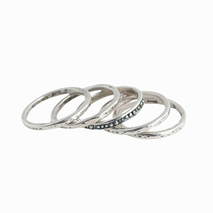 A set of five silver stackable rings with varying designs, four simple with one dotted, displayed spread out on a white background. 
