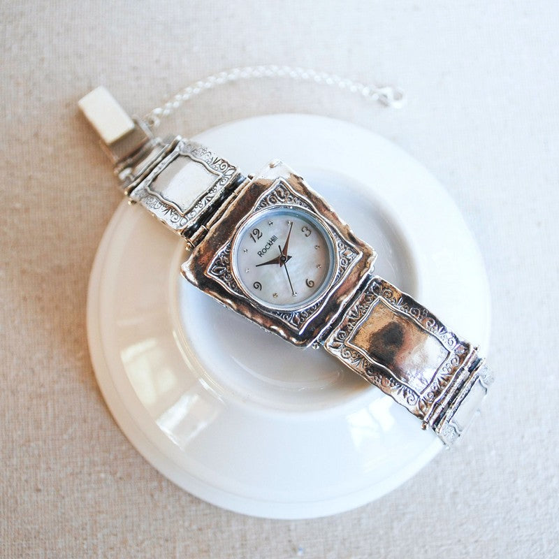 A sterling silver artisan-crafted watch with a mother-of-pearl face is shown on a white ceramic piece. 