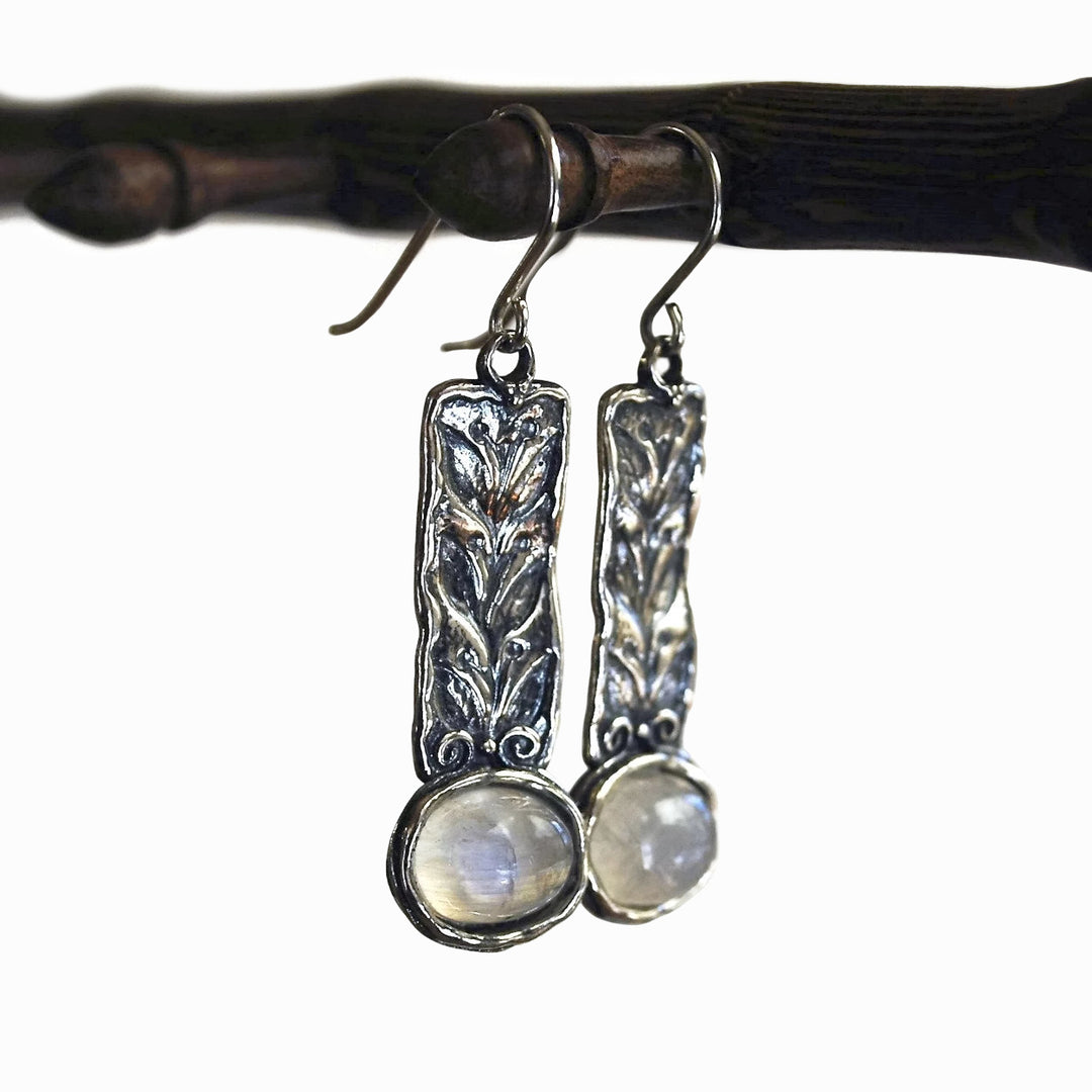 A pair of sterling silver earrings featuring rainbow moonstone cabochons and intricate leaf details, displayed dangling from a wooden dowel on a white background. 