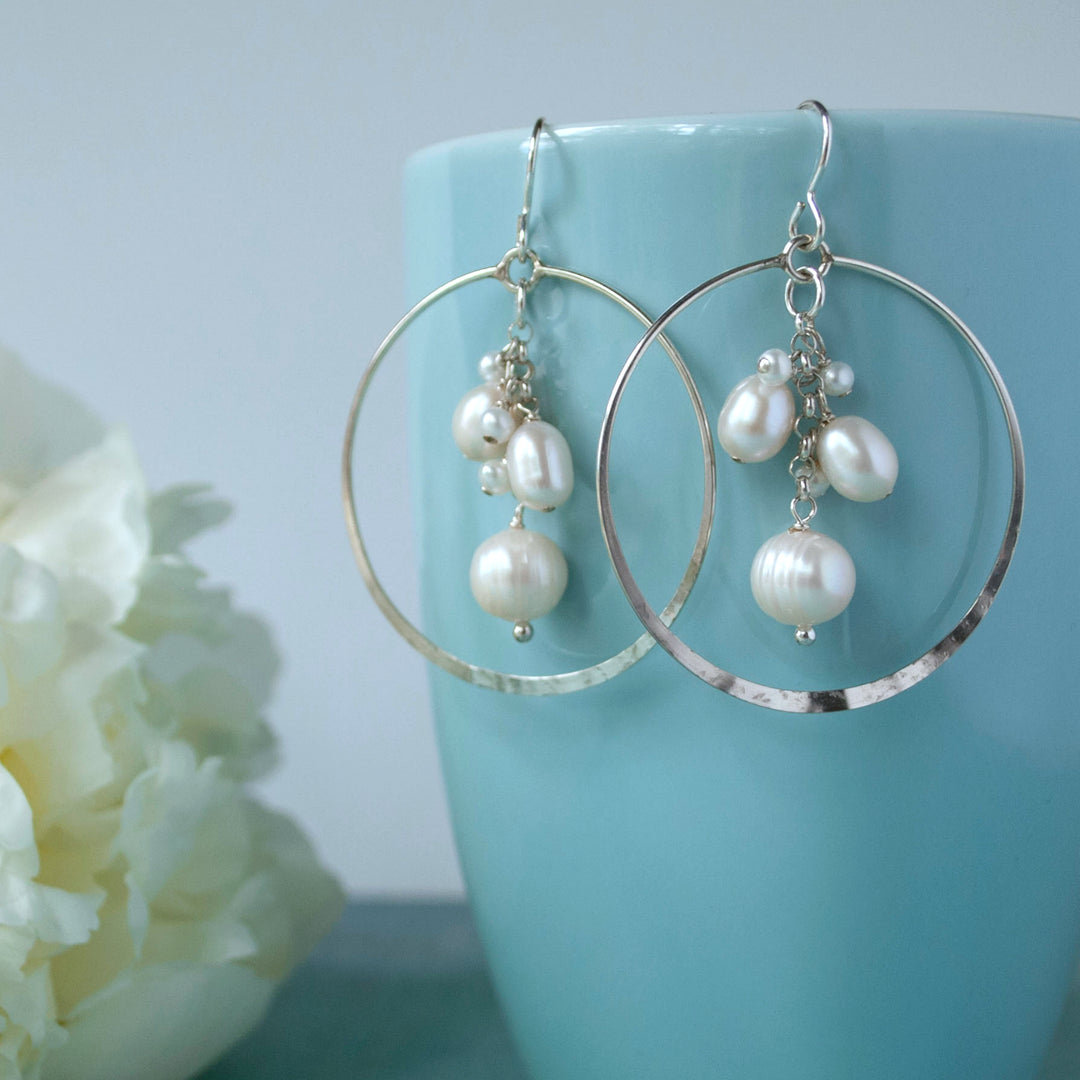 A pair of sterling silver hoop earrings with freshwater pearls attached, displayed against a blue mug with a flower.