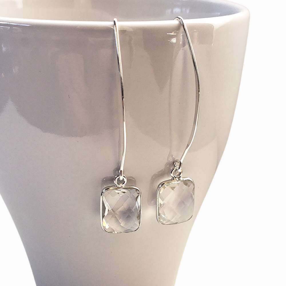Faceted clear quartz are bezel-set in sterling silver and dangle from long ear wires and are shown close up on a white mug. Earrings measure 2 1/4" long. 
