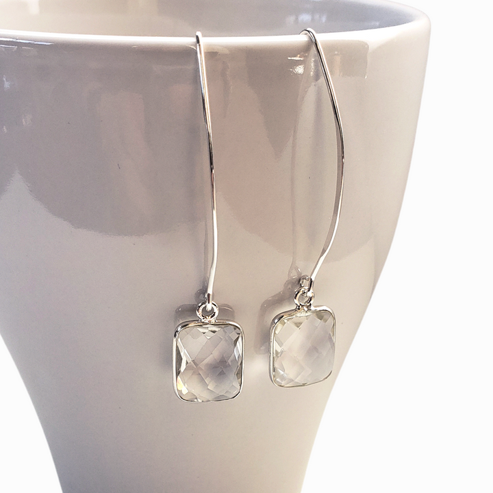 Faceted clear quartz are bezel-set in sterling silver and dangle from long ear wires and are shown close up on a white mug. Earrings measure 2 1/4" long. 