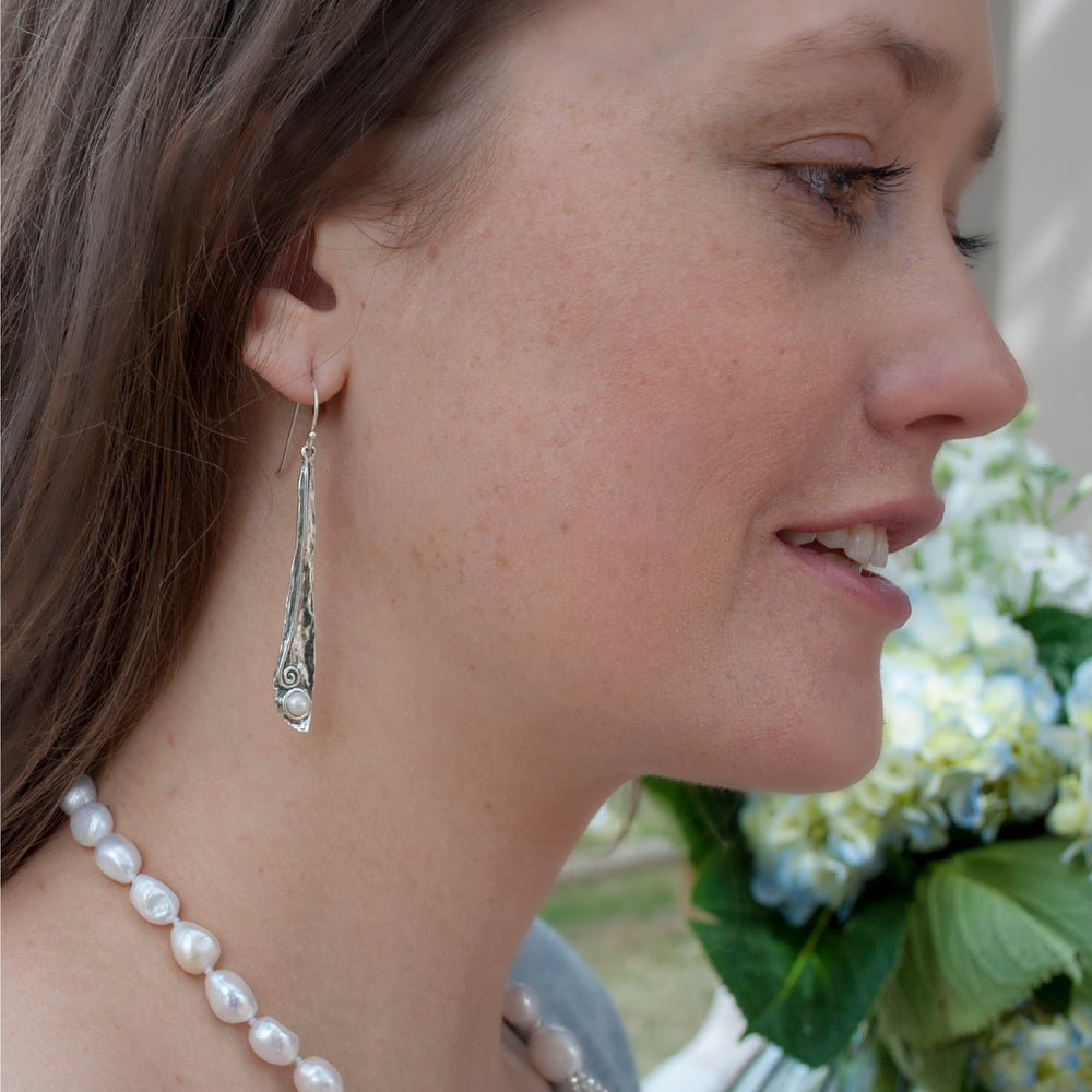 A pair of sterling silver drop earrings with a single pearl on each, attached to French earwires, shown on a model. 