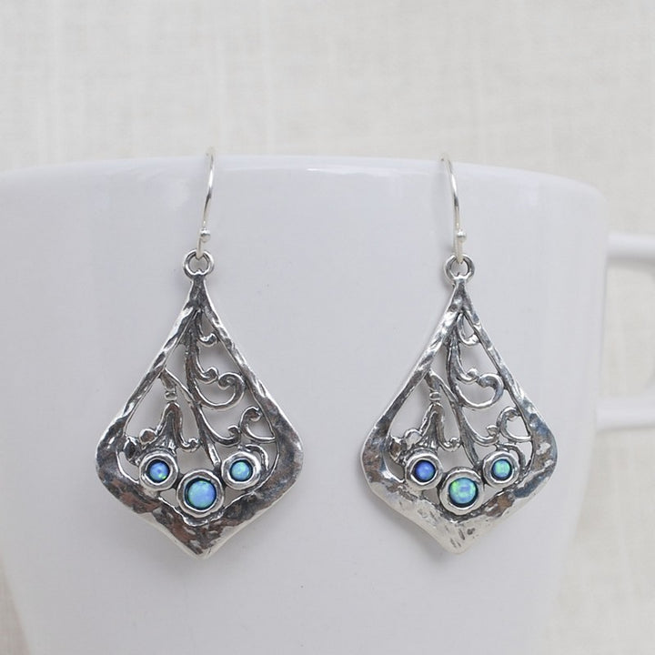 Trio of lab created blue opal is bezel set in each sterling silver earring. They are artisancrafted with scroll details. Dangle from a French ear wire. Shown on a white cup