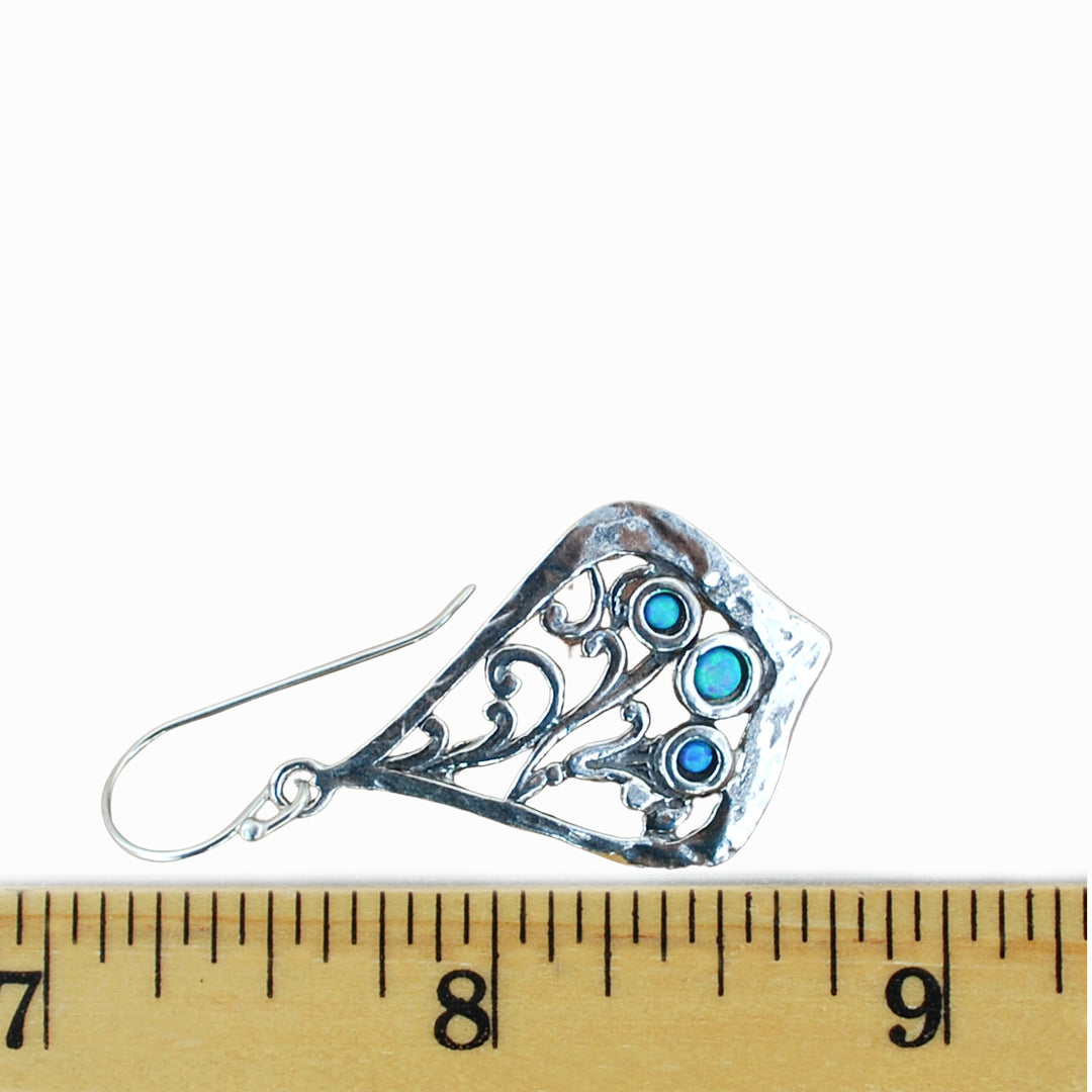 A trio of lab-created blue opals is bezel-set in each sterling silver earring. They are artisan-crafted with scroll details. Dangle from a French ear wire. Shown by a ruler with a white background for scale. 