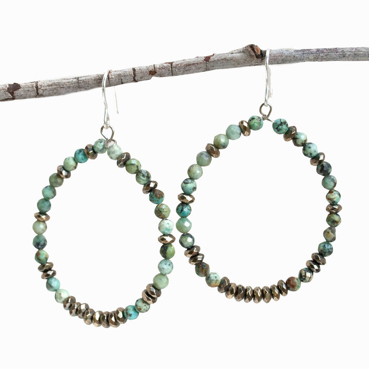 A pair of turquoise and hematite stone beaded hoop earrings with sterling silver French ear wires, displayed on a wooden branch.