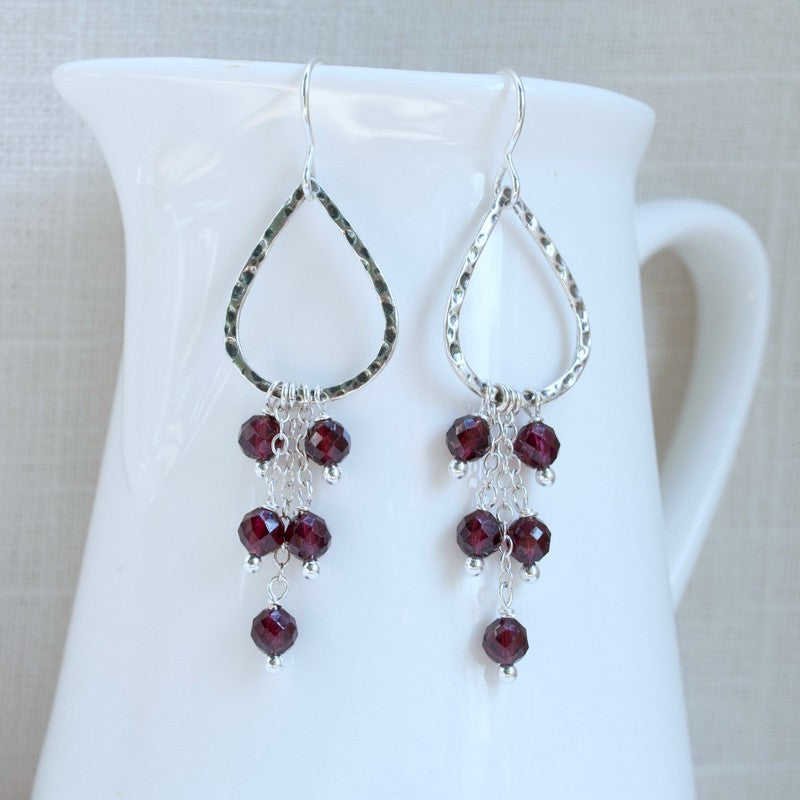 A pair of oxidized sterling silver earrings with teardrop shapes and faceted garnet stones hanging from them. Displayed dangling on a white ceramic piece. 