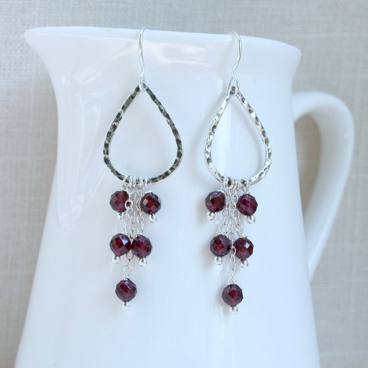 A pair of oxidized sterling silver earrings with teardrop shapes and faceted garnet stones hanging from them. Displayed dangling on a white ceramic piece. 