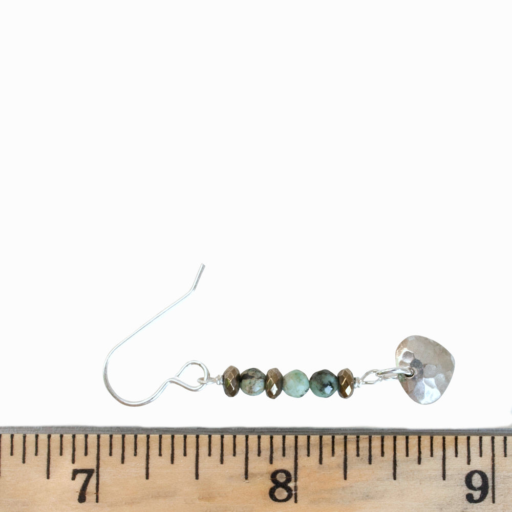 A pair of dangle earrings with stacked African turquoise and hematite beads, topped with a hammered sterling silver drop, shown by a ruler with a white background for scale. 