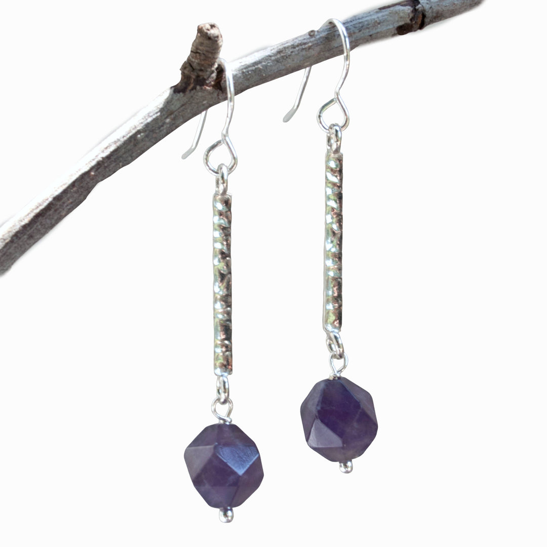 A pair of cut amethyst beads dangle from hammered sterling silver bars. Shown on a branch with a white background. 